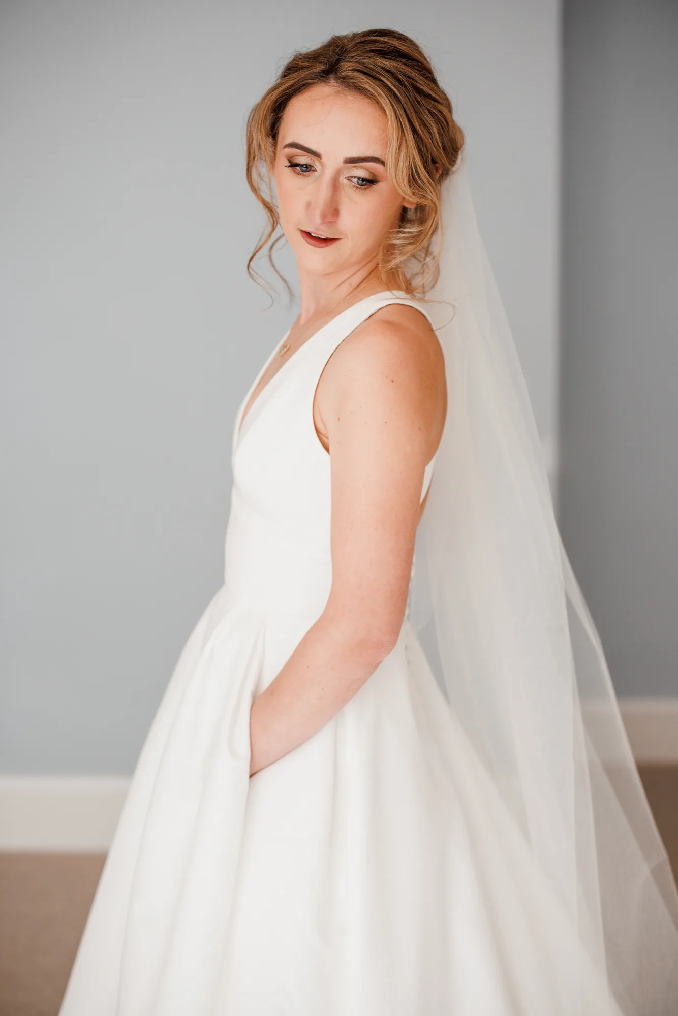 A bride in a sleeveless gown and long veil looks over her shoulder, wearing soft bronzed bridal makeup