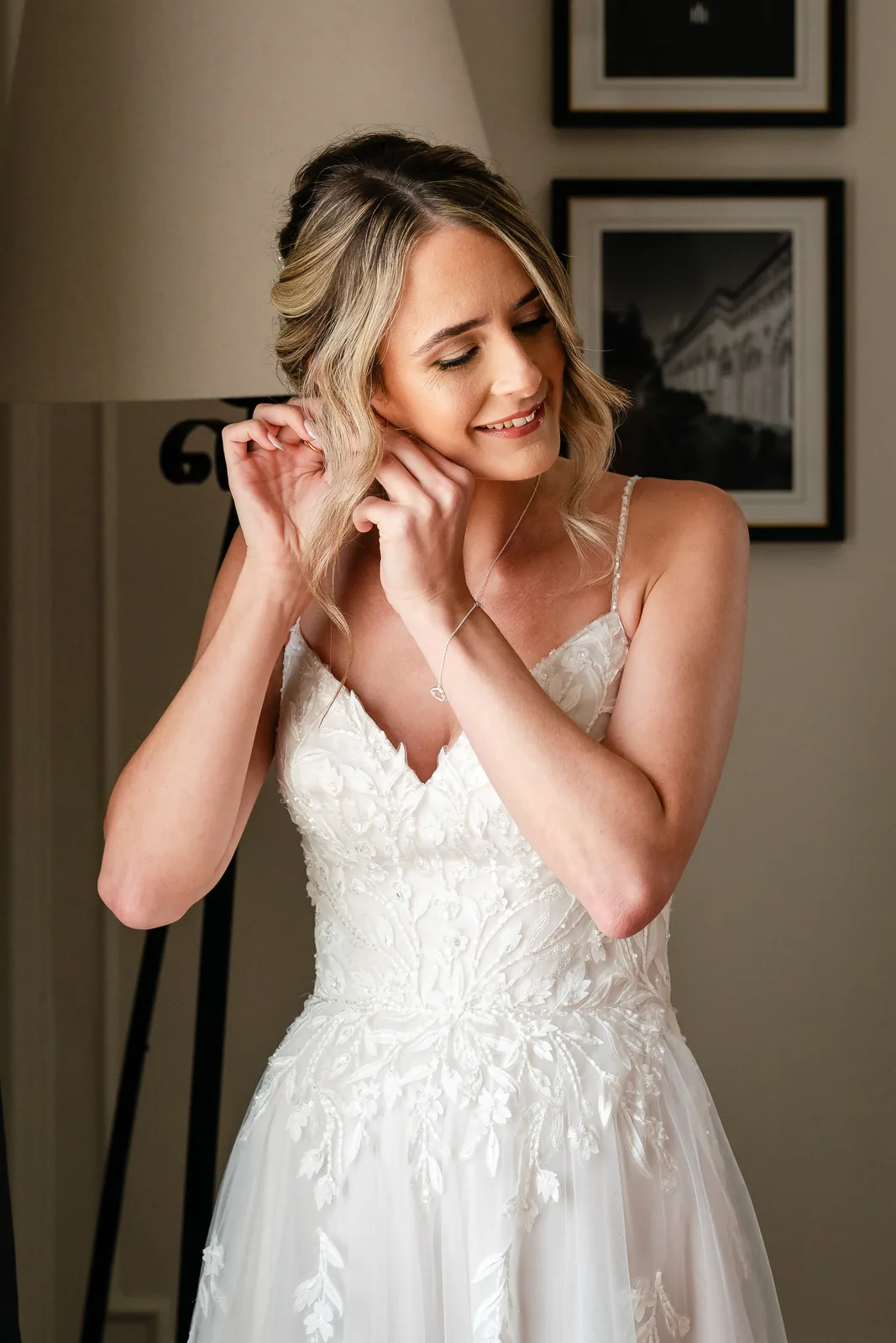 A bride in a lace gown fastening her earrings, wearing soft bridal makeup