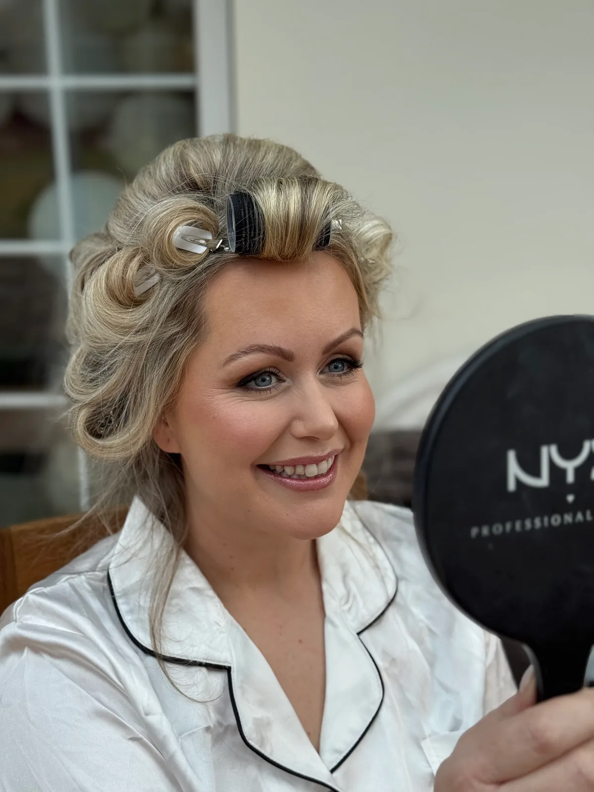 A bride in hair rollers smiling into a hand mirror as she checks her makeup
