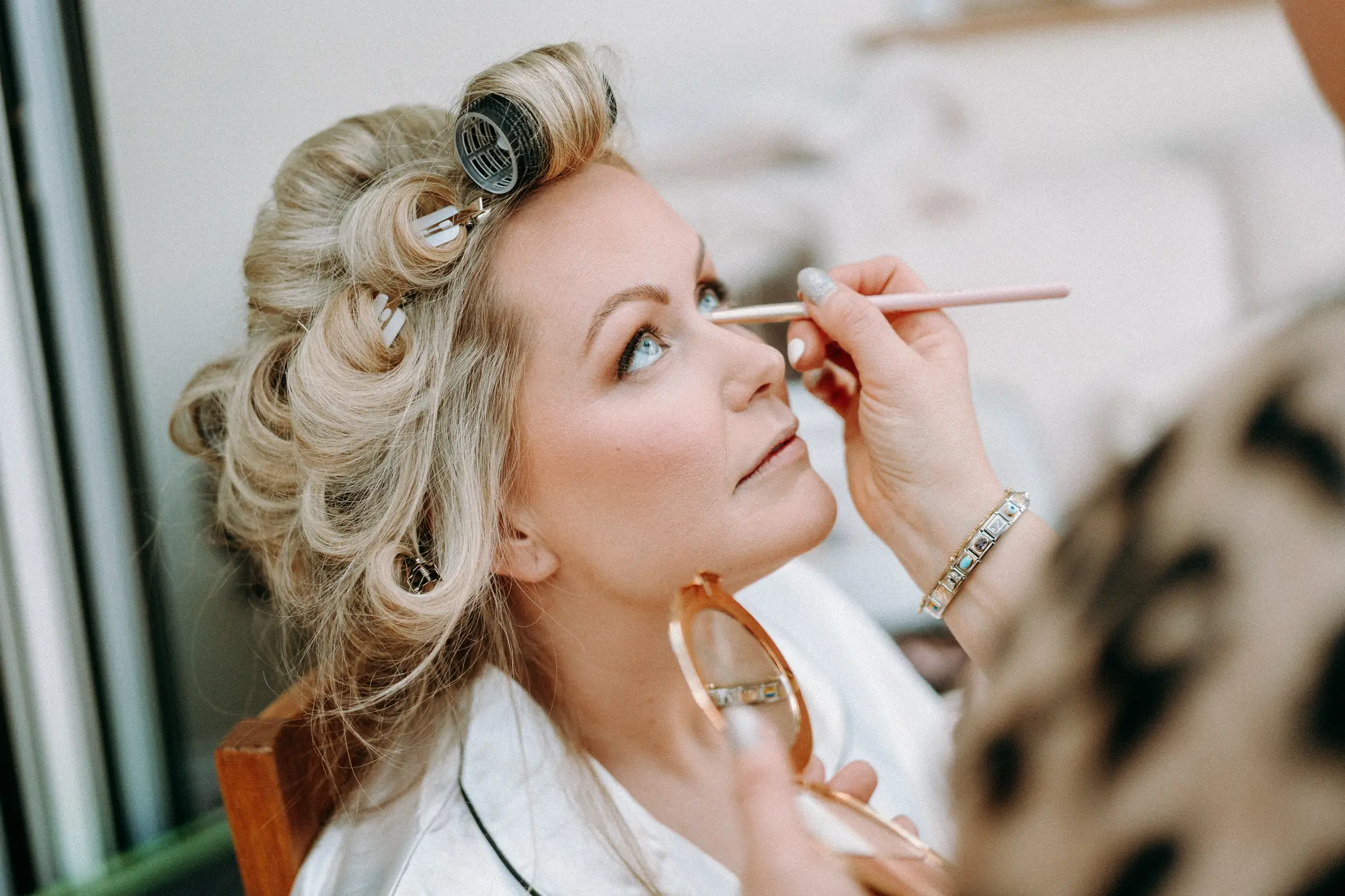 Anna applying eyeliner beneath a client's lashes, the client's hair set in rollers