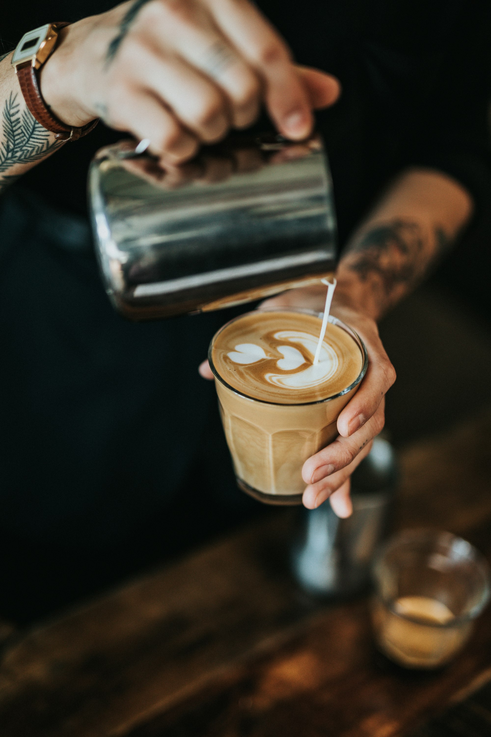 A barista pours latte art into a glass of coffee over a dark wooden counter