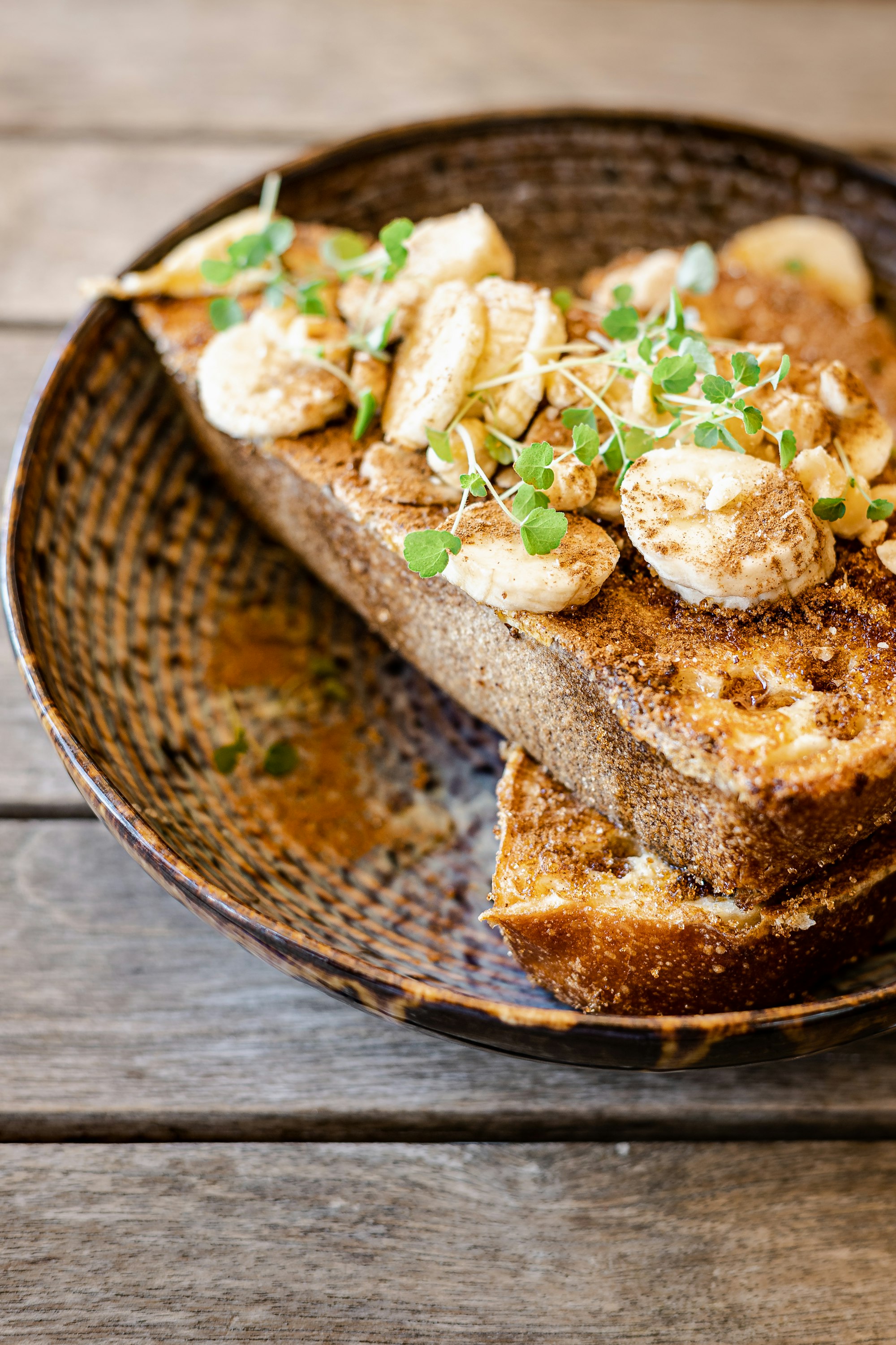 Thick toast topped with banana, cinnamon and micro-herbs in a woven bowl on a wooden table