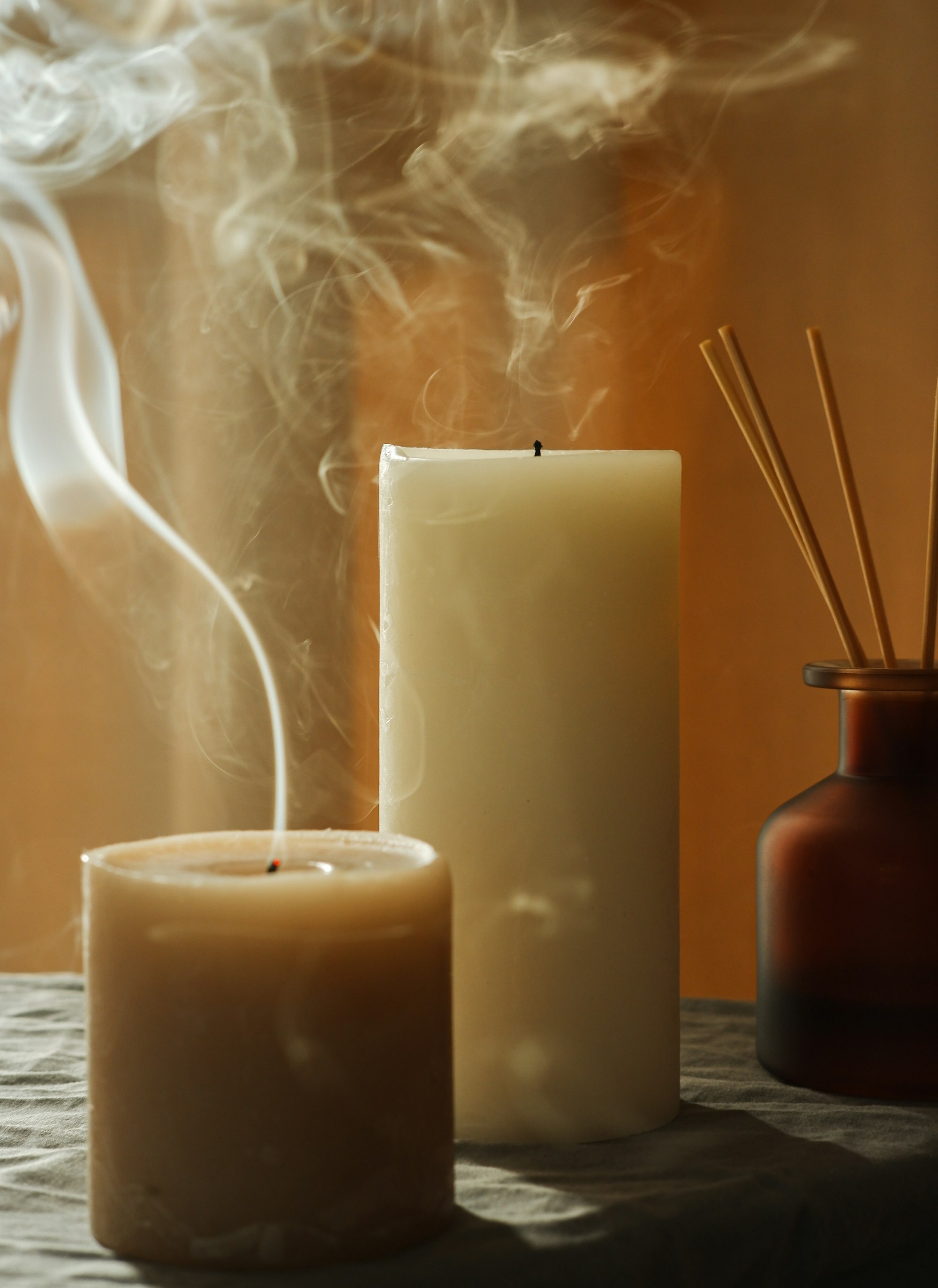 Smoke curling from a just-extinguished pillar candle beside a tall ivory pillar and an amber reed diffuser in warm light