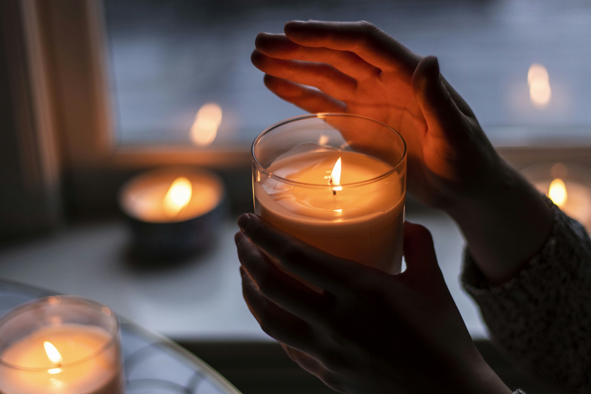 Hands cupping a lit candle in a clear glass by a window with more candles glowing behind