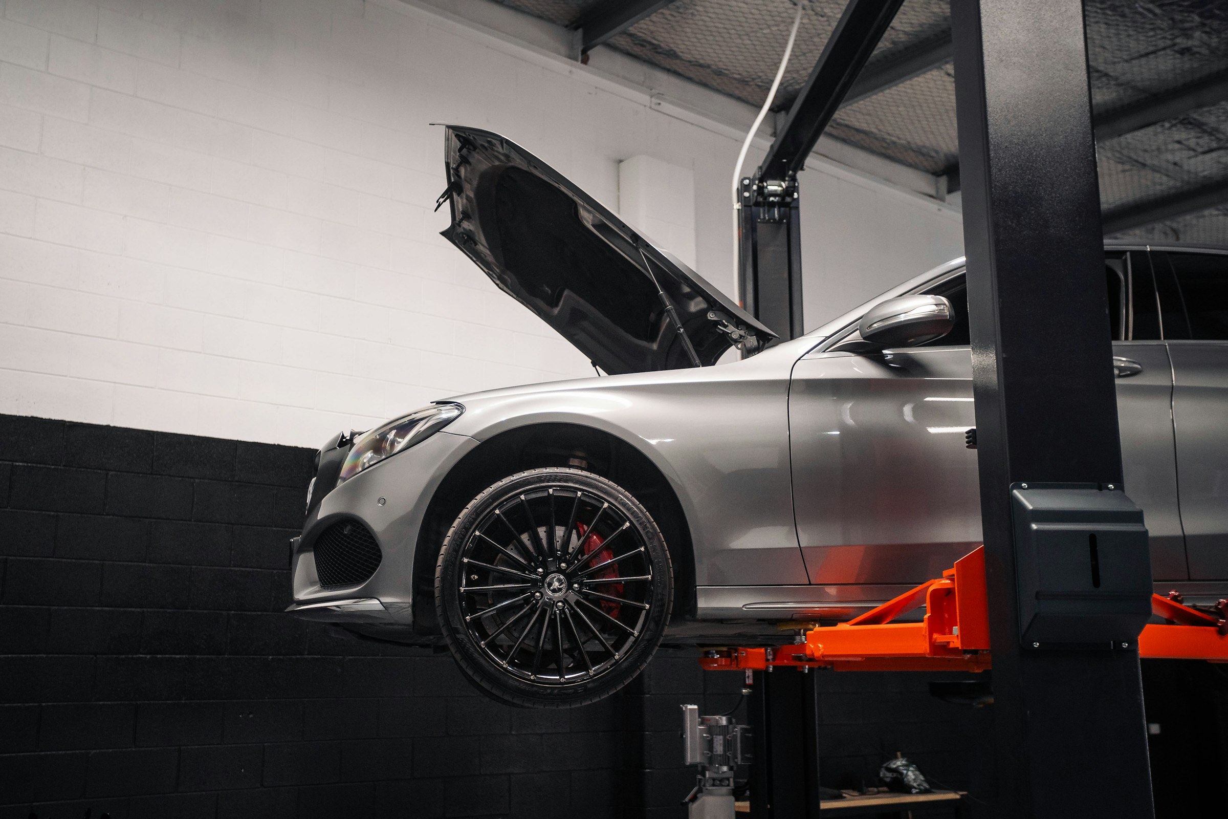 A silver saloon raised on an orange two-post lift with its bonnet up, in a white-and-black workshop bay