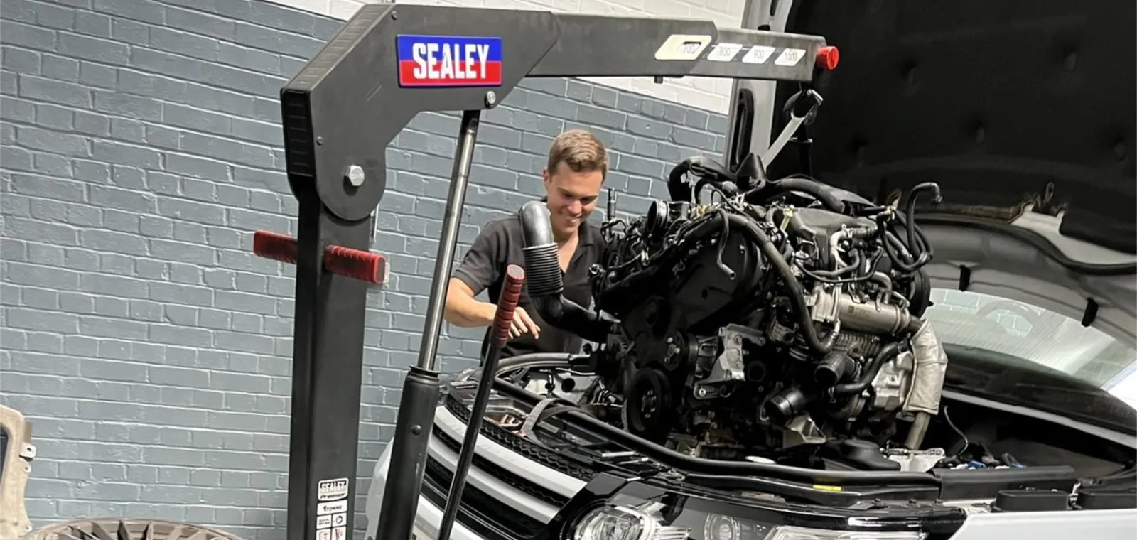 A His and Hers Motors technician lifting an engine with a workshop crane