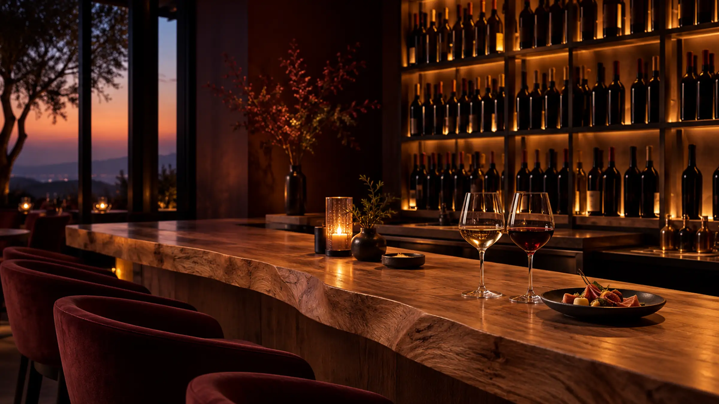 A long timber bar with two glasses of wine and a plate, lit shelves of bottles behind and a sunset through the window