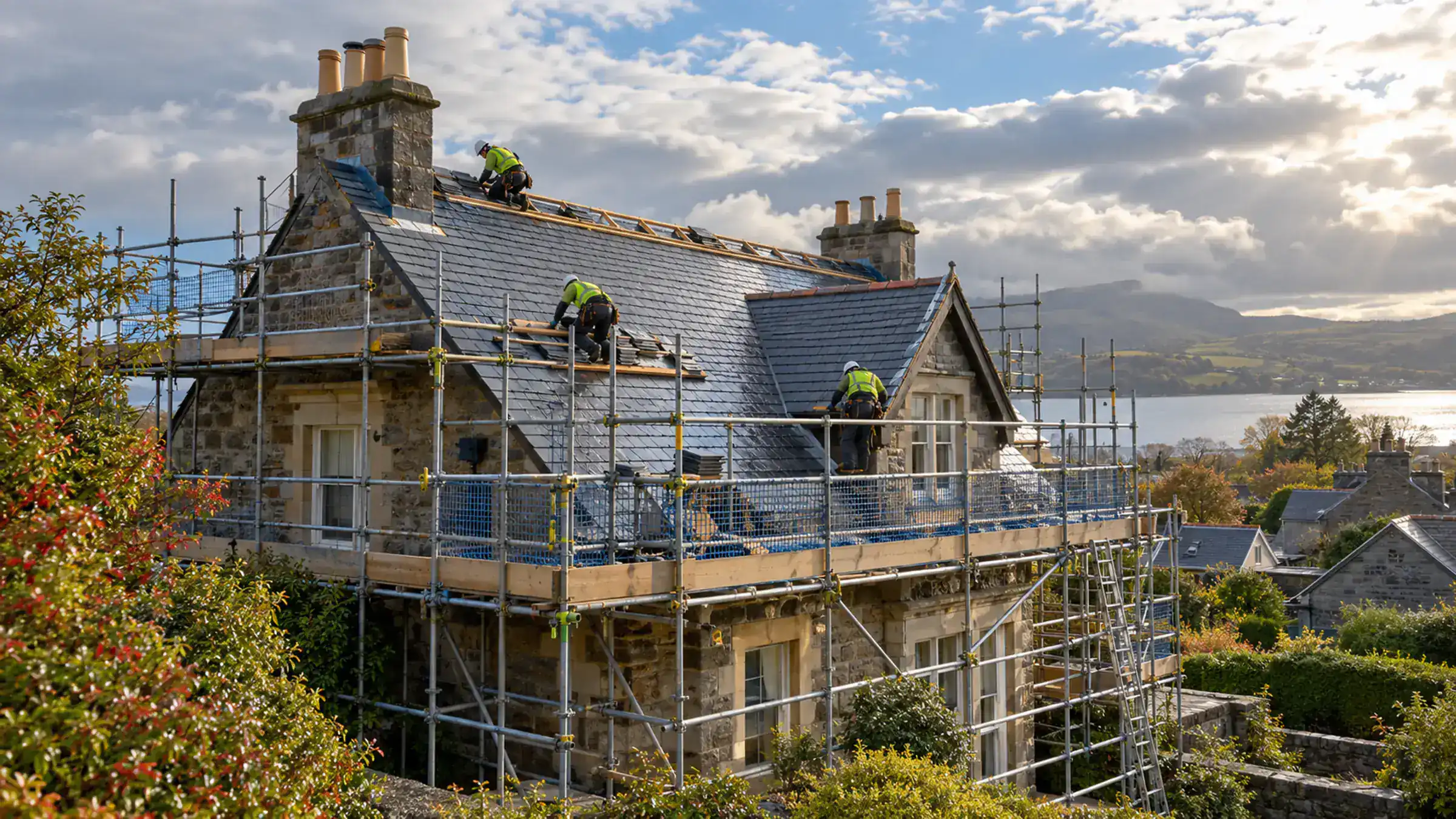 A stone house wrapped in scaffolding, two roofers working on the slate roof and hills behind