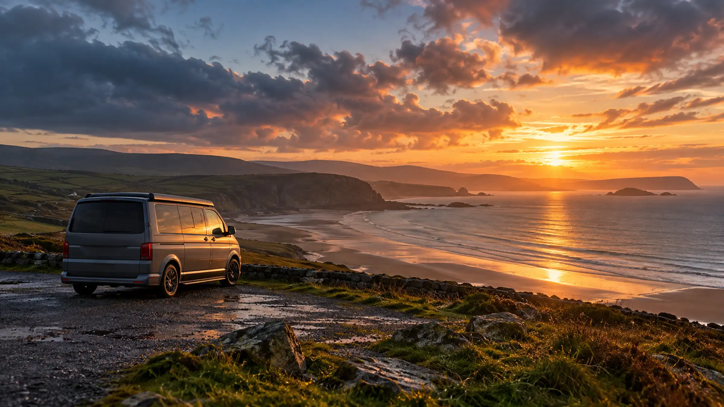 A campervan parked alone on a coastal headland at sunset, the sea and a long beach below